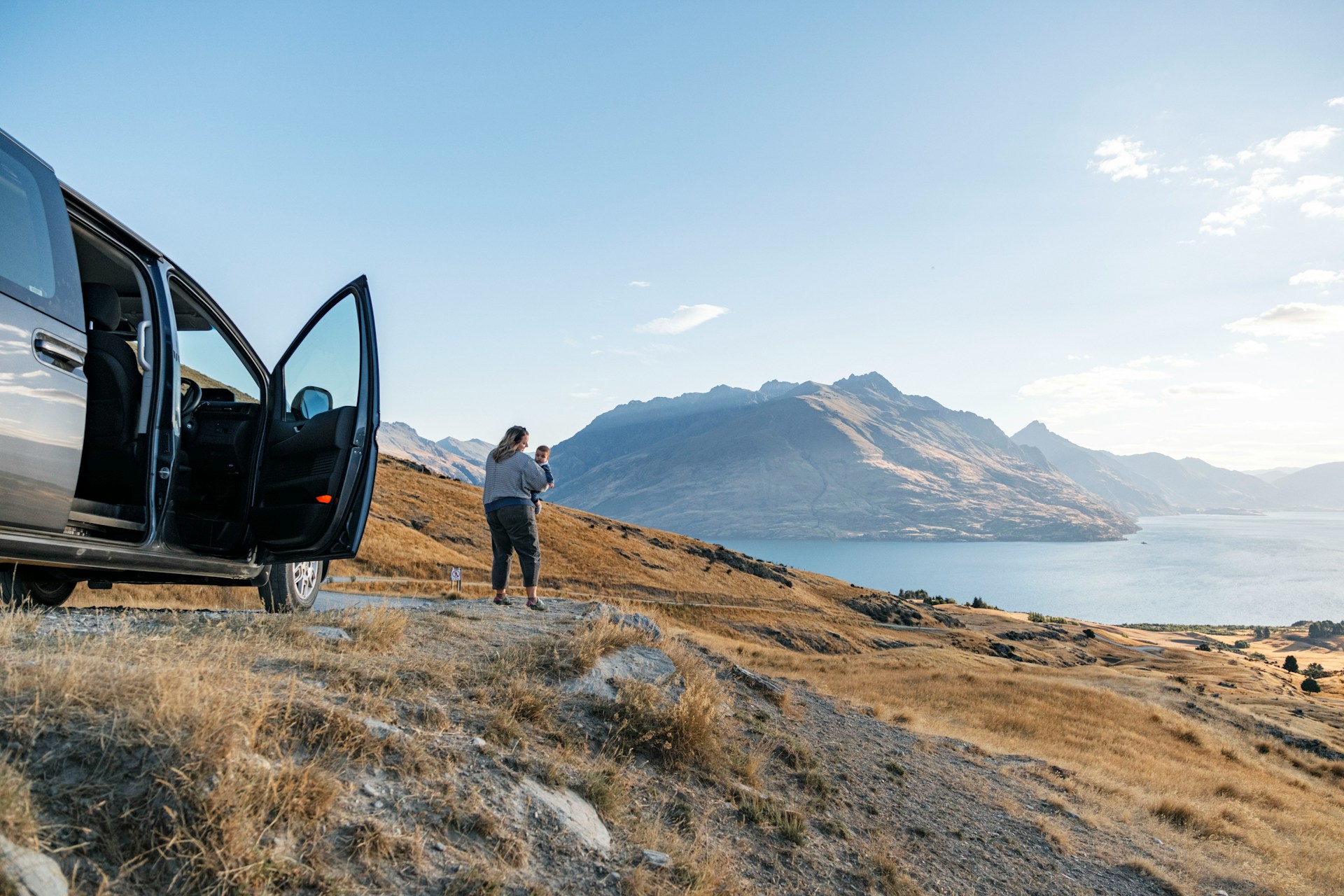 Mère portant son bébé devant un van lors d'un road trip face à un lac de montagne